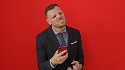 Frustrated businessman in suit frowns while holding smartphone against isolated red background