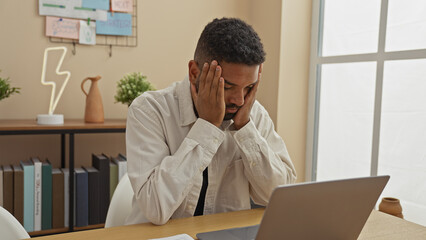 A stressed young african american man with a beard at his home office closing eyes and touching temples near laptop