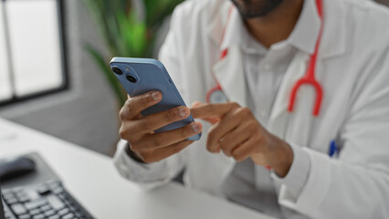 A young african american man in a white lab coat using a smartphone in a hospital office.