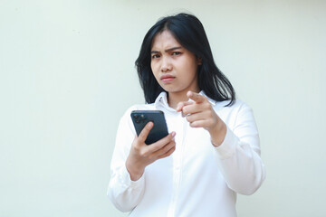Fototapeta premium portrait of sad unbelieve asian business woman pointing to camera with glare eye holding smartphone, disappointment and disatisfied expression, wear formal white suit standing over isolated background