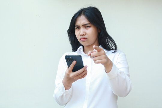 portrait of sad unbelieve asian business woman pointing to camera with glare eye holding smartphone, disappointment and disatisfied expression, wear formal white suit standing over isolated background