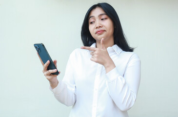 Fototapeta premium portrait of successful young asian saleswoman pointing finger with thumbs up to smartphone screen feel satisfied choose product in online shope wearing white formal suit shirt over isolated background