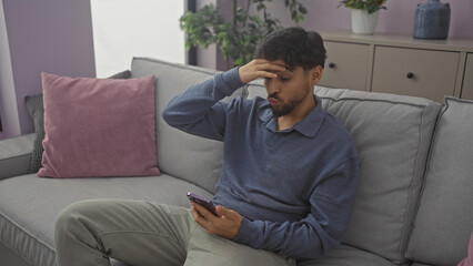 A stressed man sitting on a gray couch holding a smartphone in a modern apartment.