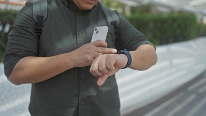 Hispanic man checks smartwatch with smartphone in hand outdoors in the city.