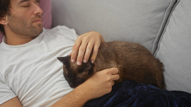 Hispanic man resting at home with his siamese cat on a couch, conveying comfort and companionship.