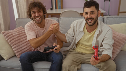 Men playing video games and laughing together on a cozy couch in a well-lit living room, shaking...