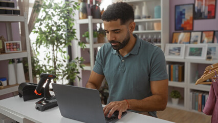Fototapeta premium Young man working on a laptop in a home decor store, surrounded by various decorations and indoor plants.