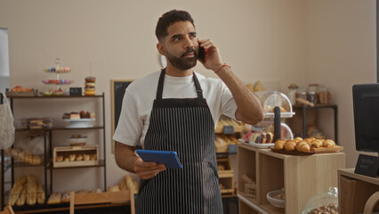 Hispanic man in bakery wearing apron, holding tablet, and talking on phone in a room filled with bread and pastries.