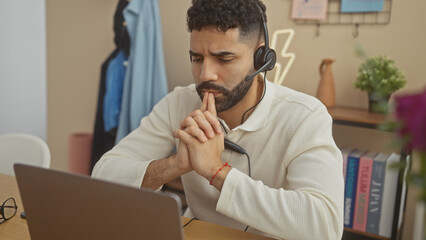 Fototapeta premium A thoughtful hispanic man with a beard wears a headset while working on a laptop in a cozy home office.