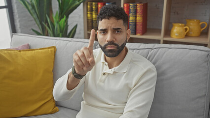 Hispanic man with beard in a white shirt sitting on a gray couch indoors raising one finger