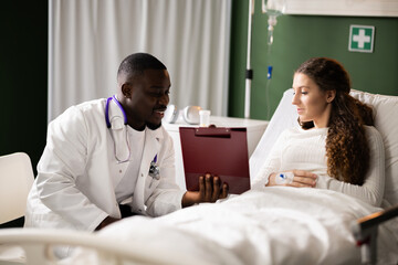 Young African doctor sits by the red-haired girl, presenting information from a folder during a consultation.