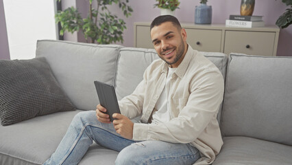 A smiling young hispanic man holding a tablet while sitting on a gray couch in a well-decorated living room