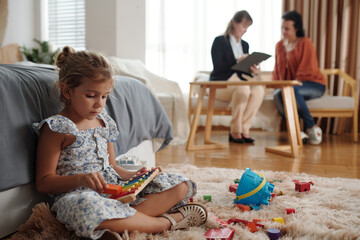 Little girl sitting on the floor and playing toys while her mom talking to social worker