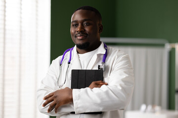 Enjoying a peaceful moment, the doctor stands in a green room, smiling and looking out the window. He holds a tablet and has a stethoscope around his neck.