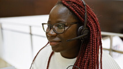 African american woman with braids wearing headset smiles in a corporate office environment.