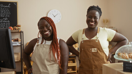 Two african american women bakers smiling in a cozy bakery shop with pastries and bread, indoor setting with a clock visible on the wall behind them.