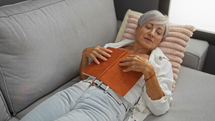 Obraz premium Elderly woman with short grey hair resting in a living room at home, holding an orange book on her chest while laying on a grey couch with a striped pink pillow