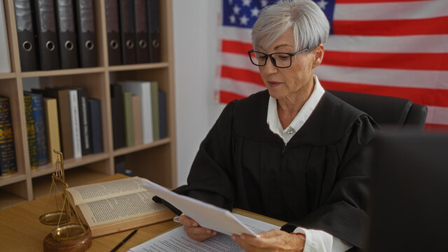 Elderly woman judge with short grey hair reading documents in a courtroom setting with american flag in background