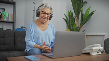 Senior woman with glasses and headphones using laptop in a cozy living room.