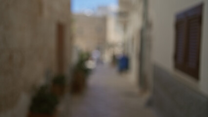 Blurry, narrow alleyway with mediterranean stone walls, plants in pots, and blue sky in the...