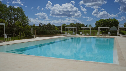 A serene outdoor pool with lush greenery and clear blue skies, surrounded by lounge chairs indicative of a peaceful, sunny day, perfect for relaxation and leisure activities.