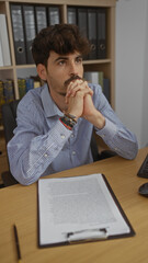 Naklejka premium Handsome young hispanic man with beard sitting in office with thoughtful expression in front of clipboard and computer
