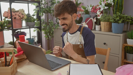 A joyful young man with a beard celebrates success in a vibrant flower shop environment, interacting with a laptop.