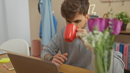 A young hispanic man sips coffee indoors at a home office with a laptop, books, and flowers in the...