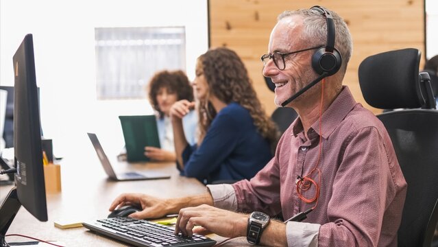 Happy mature businessman using computer and headset in coworking office - Powered by Adobe