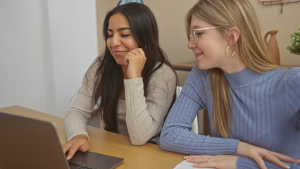 Fototapeta premium Two women friends are smiling and working together on a laptop in a modern home interior.