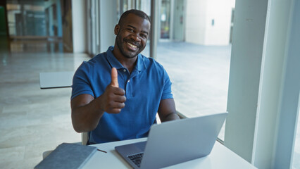 Attractive young man in a workplace giving a thumbs up while working on a laptop indoors.