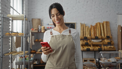 Woman smiling while holding a cellphone in a bakery with fresh bread and pastries on display