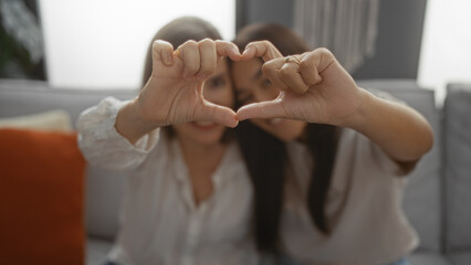 Mother and daughter forming heart with their hands, sitting together in a cozy living room,...