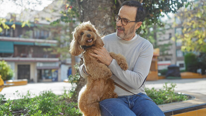 A middle-aged hispanic man enjoys outdoor time in an urban park with his fluffy poodle, against a...