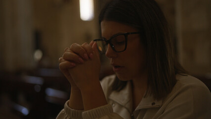 A young, beautiful hispanic woman praying with closed eyes and clasped hands inside a historic church in italy, capturing the somber and devout christian atmosphere.