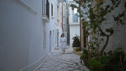 Fototapeta premium A young hispanic woman in stylish white attire walks through a charming narrow street in polignano a mare, puglia, italy, surrounded by white-washed walls and lush greenery.