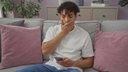 A concerned hispanic man in a white shirt sitting on a gray sofa at home, holding a phone and...