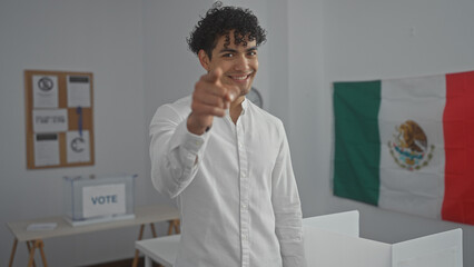 Smiling young hispanic man pointing, wearing a white shirt in a voting room with a mexican flag in...
