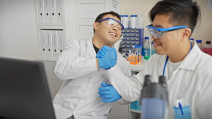Two smiling men in lab coats and safety glasses giving a friendly fist bump in a laboratory setting.