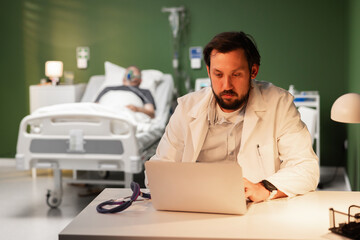 A middle-aged doctor sits tiredly at his desk in a hospital room. An overtired doctor at work © ABCreative