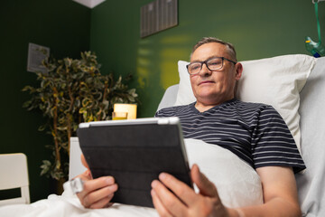 Resting in his hospital bed, the elderly man recuperates while enjoying his tablet, engrossed in heartwarming messages from loved ones, fueling his determination to heal.