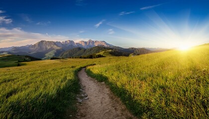 Obraz premium inviting dirt trail winds through a lush green field towards a stunning mountain range illuminated by the golden light of the afternoon sun
