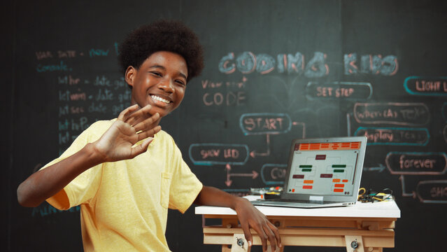 Smart african boy use laptop to code program and turn around to wave hand. Cute highschool stident coding engineering prompt or program system at blackboard in STEM technology classroom. Edification