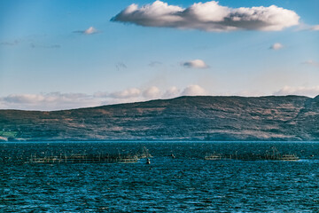 The view shows a vast expanse of still water with few fish farms positioned in the centre, against the backdrop of distant dark mountains.