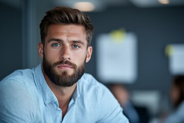 Obraz premium A bearded man in a blue shirt looks confidently at the camera, capturing a moment of introspection and determination in a contemporary office environment.