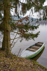 Obraz premium Old wooden boat on water near pine tree on bank of calm Vltava river, Slapy, Czech Republic, winter.