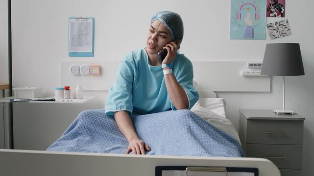 Young female patient in hospital gown and cap with pre-surgical marks on her face sitting on bed in medical room and talking on mobile phone while preparing for plastic surgery in clinic