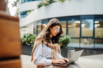 Young businesswoman working on laptop outside of modern business building