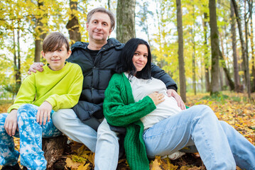 Happy family in autumn park. Father, pregnant mother and son sitting on fallen leaves.