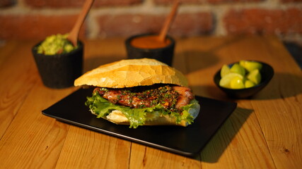 Choripan con chimichurri servido en un plato negro acompañado de salsa roja y verde, guacamole  y limones